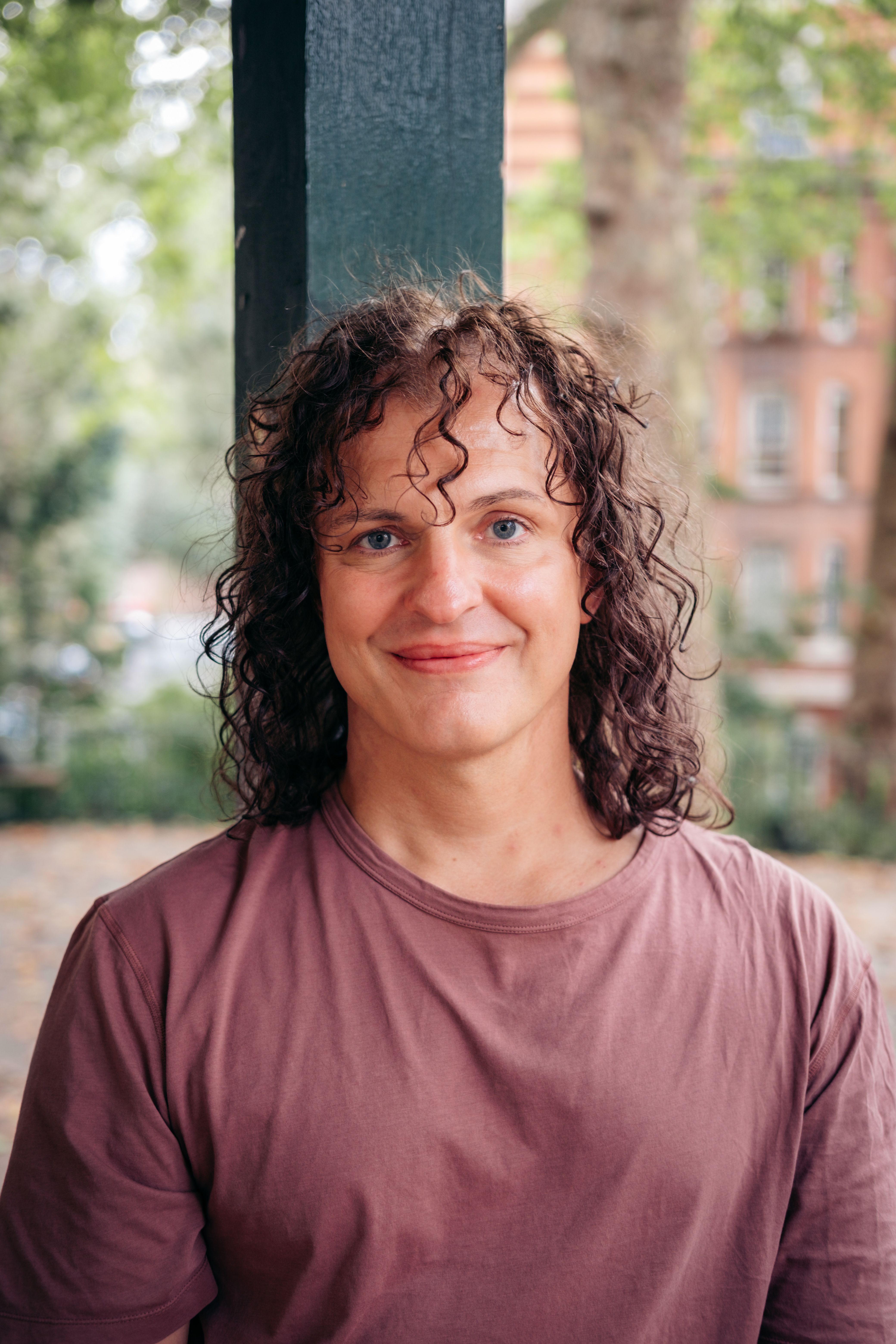 Headshot of Gabriel Muller-Ebeid, a man with long curly brown hair and blue eyes, smiling warmly at the camera in a park.